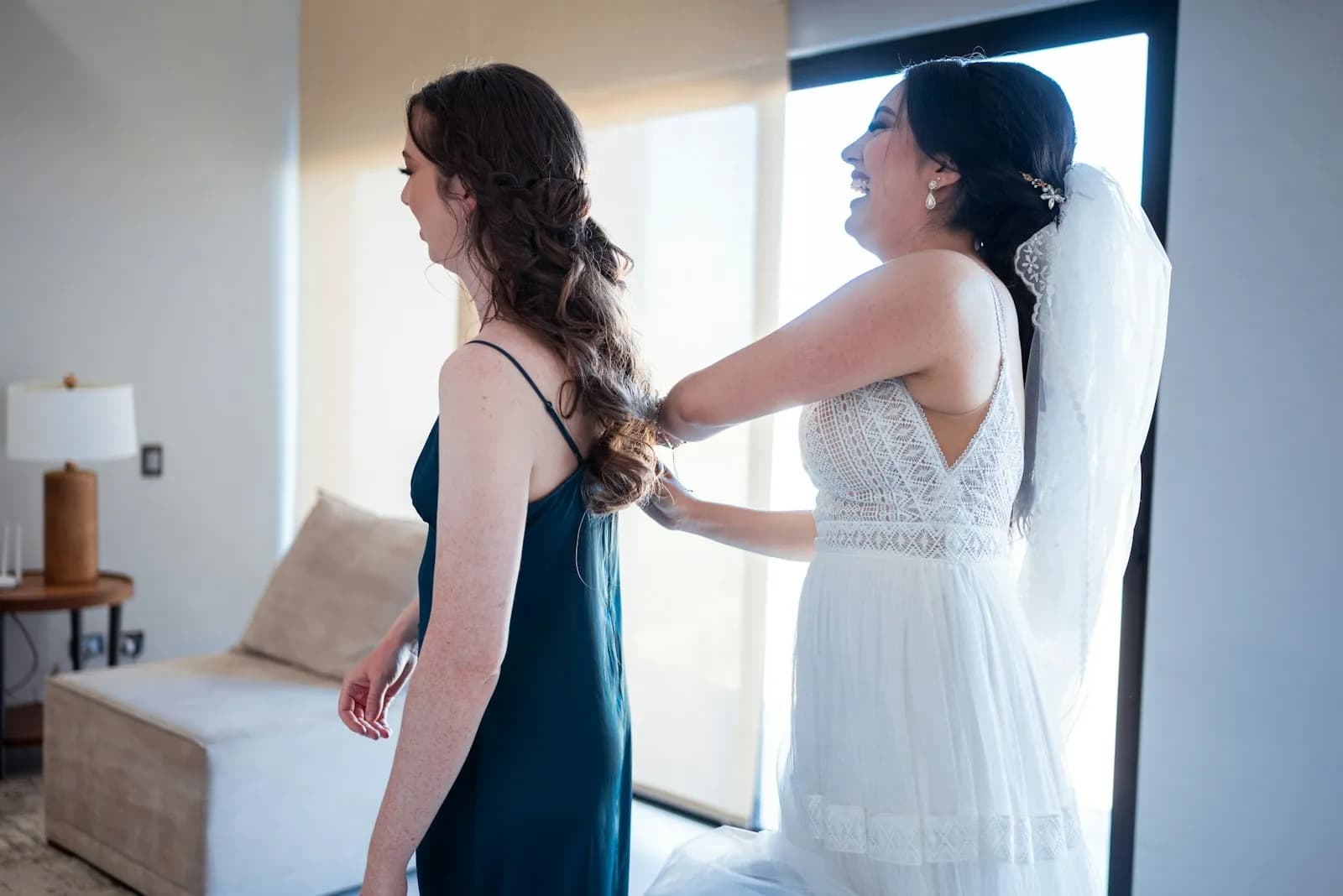 A bride in her veil laughing as she fixes a bridesmaid's hair by a bright window.