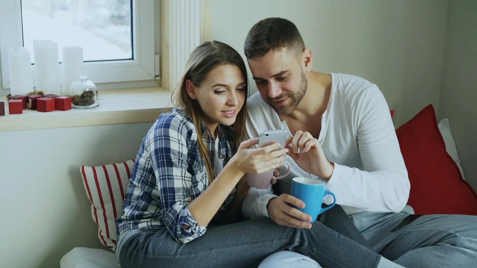A couple on a window seat looking at a phone together, smiling, with mugs of coffee.